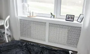 Elegancka osłona kaloryfera z ażurowym wzorem, która stanowi estetyczne rozwiązanie, jak zabudować kaloryfer. Na parapecie zdjęcia i ozdoby.