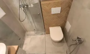 Kompaktowa, bardzo mała łazienka z prysznicem. W rogu biała miska WC, obok niej panel spłuczki z dwoma przyciskami. Ściana za WC ozdobiona drewnianymi płytkami w jodełkę.