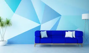 Niebieska sofa stoi przed ścianą pomalowaną do połowy w geometryczne wzory. Obok stoi lampa i roślina.