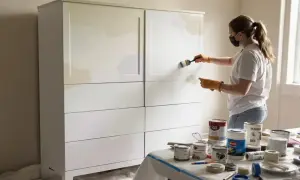 Kobieta w masce i rękawiczkach maluje białą szafę na jasny kolor. Widać puszki z farbą i taśmę malarską.