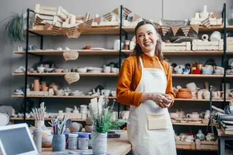Uśmiechnięta artystka w fartuchu w pracowni ceramicznej, otoczona półkami z wyrobami. Zastanawia się, jak wycenić rękodzieło, by docenić swój trud.