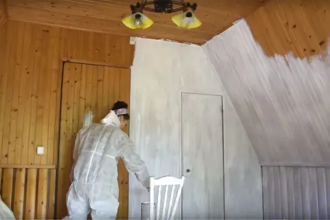 Malowanie lakierowanej boazerii bez szlifowania. Kobieta w kombinezonie maluje ściany na biało.