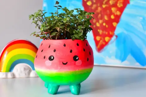 Urocza doniczka diy w kształcie arbuza z uśmiechniętą buźką, pełna zielonych roślinek. Obok tęczowa ozdoba.