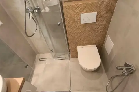 Kompaktowa, bardzo mała łazienka z prysznicem. W rogu biała miska WC, obok niej panel spłuczki z dwoma przyciskami. Ściana za WC ozdobiona drewnianymi płytkami w jodełkę.