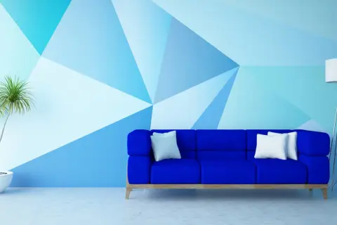 Niebieska sofa stoi przed ścianą pomalowaną do połowy w geometryczne wzory. Obok stoi lampa i roślina.
