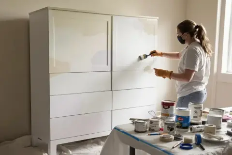 Kobieta w masce i rękawiczkach maluje białą szafę na jasny kolor. Widać puszki z farbą i taśmę malarską.