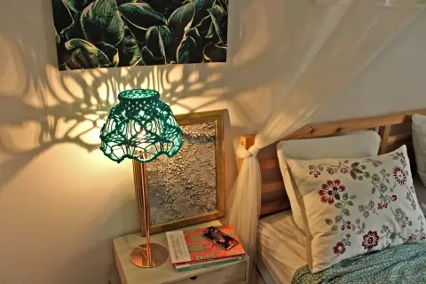 Zielona, pleciona lampa nocna rzuca cienie na ścianę. Obok łóżko z poduszką.