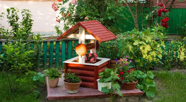 Uroczy, drewniany domek studni z ozdobnymi detalami, otoczony kwitnącymi roślinami. Ciekawe ozdoby z drewna do ogrodu dodają mu charakteru.