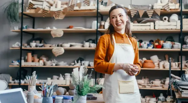 Uśmiechnięta artystka w fartuchu w pracowni ceramicznej, otoczona półkami z wyrobami. Zastanawia się, jak wycenić rękodzieło, by docenić swój trud.