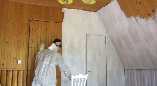 Malowanie lakierowanej boazerii bez szlifowania. Kobieta w kombinezonie maluje ściany na biało.