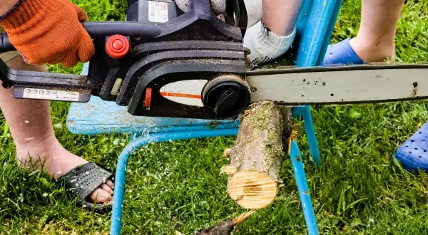 Osoba w pomarańczowej rękawicy używa piły łańcuchowej do cięcia drewna na prowizorycznym stojaku. To prosty sposób, jak zrobić stojak do cięcia drewna.