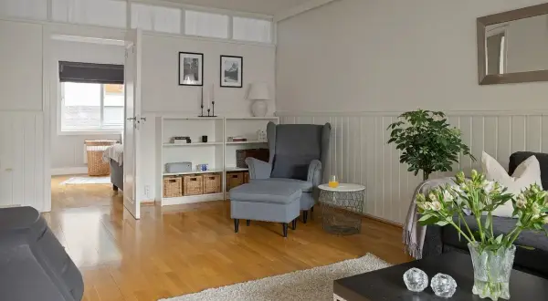 Jasny salon z pomalowaną boazerią, szarym fotelem i kanapą, książkami i wiklinowymi koszami.