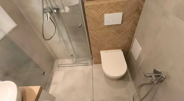 Kompaktowa, bardzo mała łazienka z prysznicem. W rogu biała miska WC, obok niej panel spłuczki z dwoma przyciskami. Ściana za WC ozdobiona drewnianymi płytkami w jodełkę.
