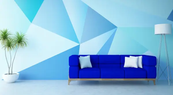 Niebieska sofa stoi przed ścianą pomalowaną do połowy w geometryczne wzory. Obok stoi lampa i roślina.