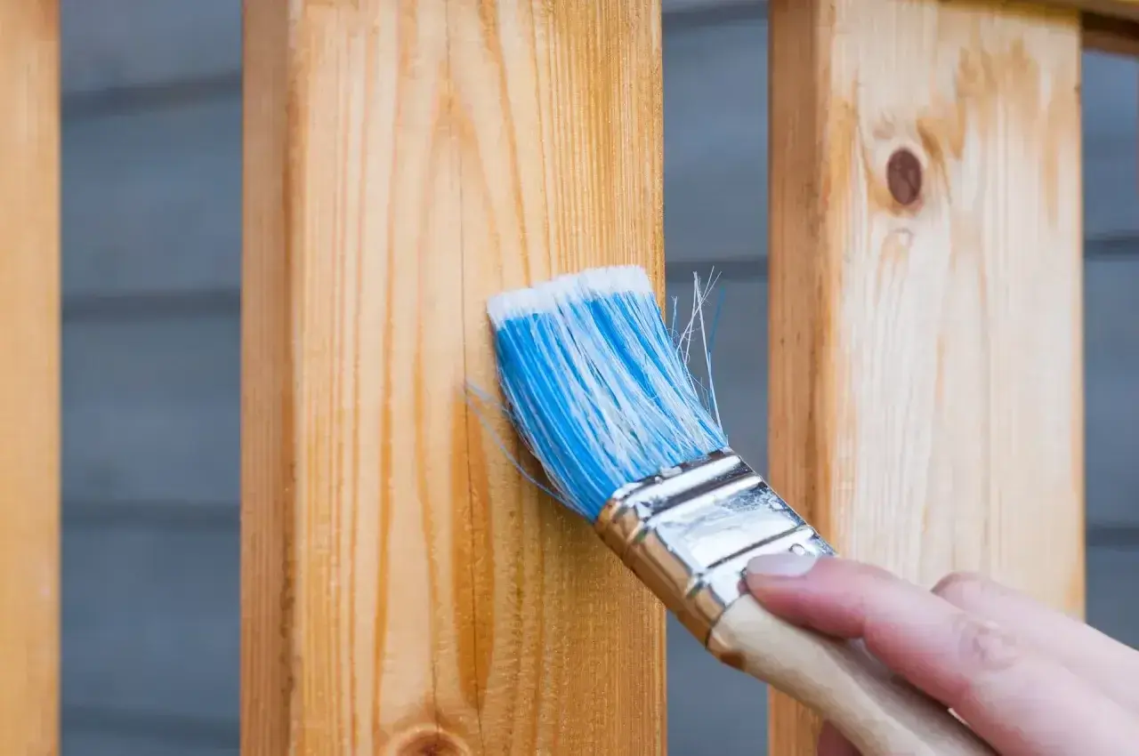 Pędzel z niebieskim włosiem nakłada bezbarwny impregnat na drewniane balustrady. Dowiedz się, czym zabezpieczyć drewno na zewnątrz, by służyło latami.