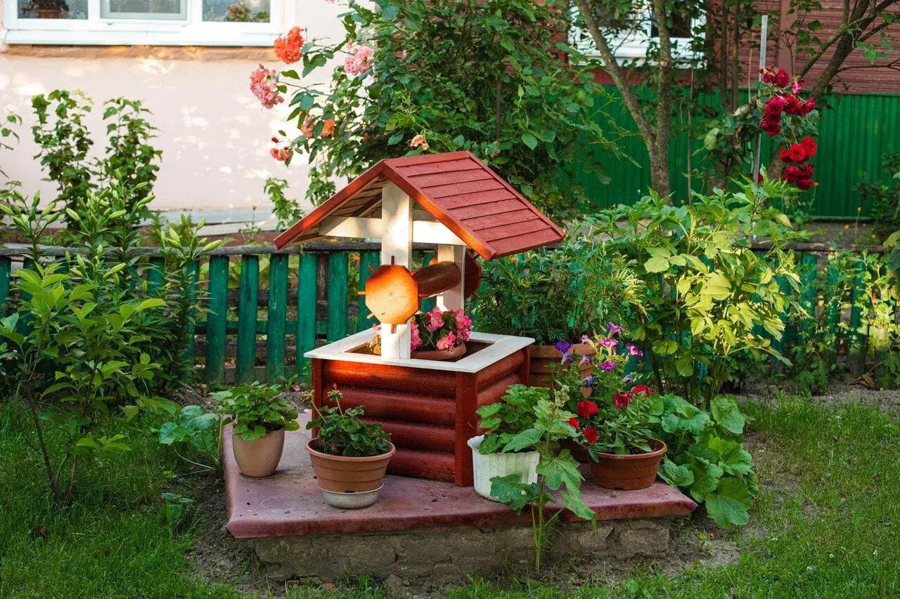 Uroczy, drewniany domek studni z ozdobnymi detalami, otoczony kwitnącymi roślinami. Ciekawe ozdoby z drewna do ogrodu dodają mu charakteru.