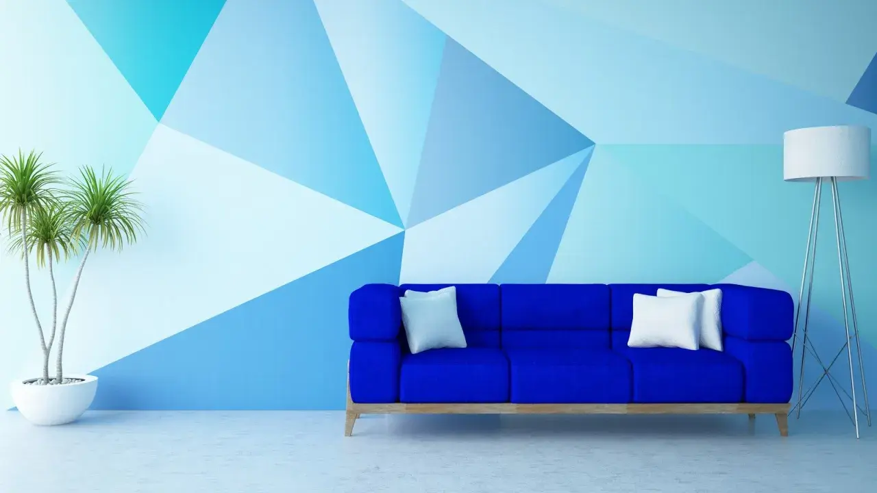 Niebieska sofa stoi przed ścianą pomalowaną do połowy w geometryczne wzory. Obok stoi lampa i roślina.