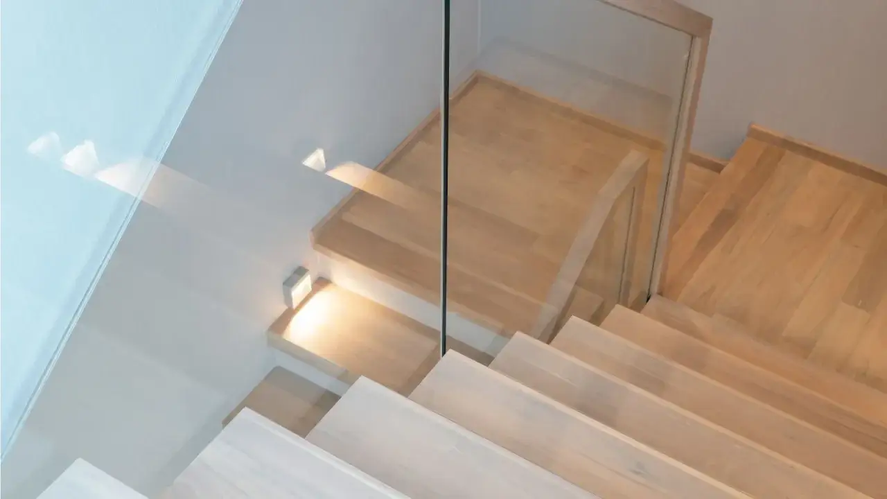 Nowoczesne schody drewniane z jasnym lakierem, szklaną balustradą i dyskretnym oświetleniem.