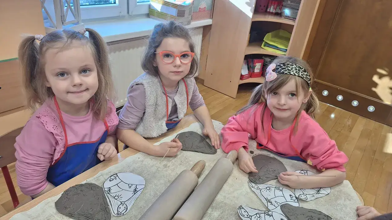 Trzy dziewczynki tworzą dzieła z gliny podczas warsztatów. Na stole leżą wałki i gliniane formy.