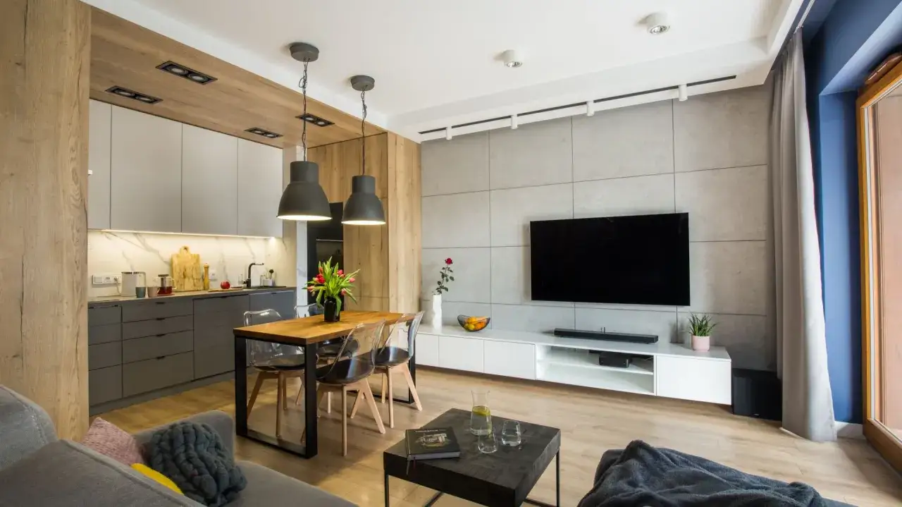 Nowoczesny salon z kuchnią. Beton i drewno tworzą przytulną atmosferę. W centrum drewniany stół z przezroczystymi krzesłami.