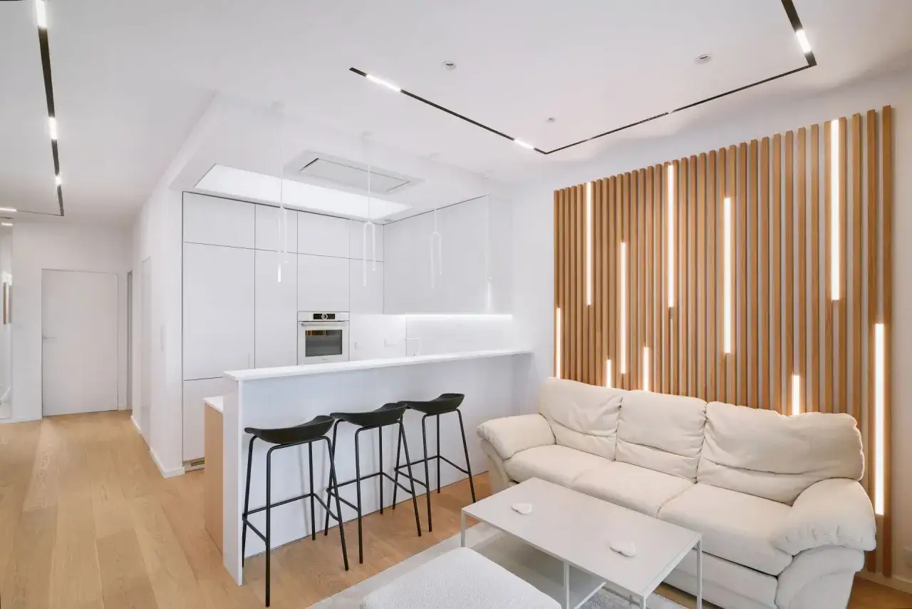 Nowoczesny salon w drewnie i bieli. Jasna kanapa, drewniana ściana z podświetleniem i minimalistyczna kuchnia tworzą przytulną atmosferę.
